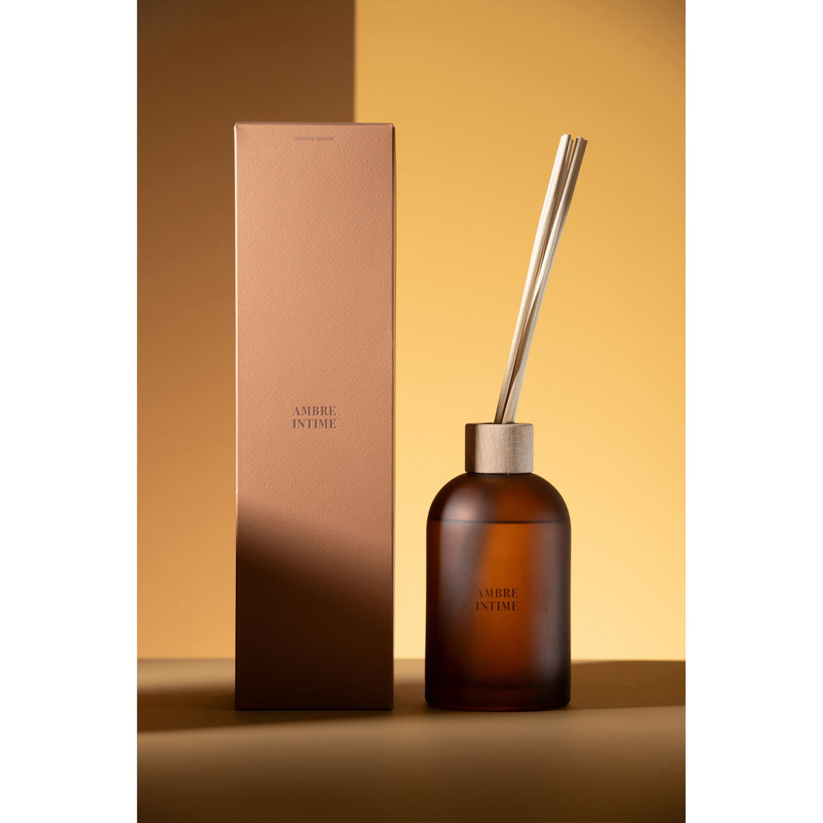 Geurstokjes Accords Essentiels Ambre Intime 550ml van J-Line gestyled tegen een okerkleurige wand met zacht licht